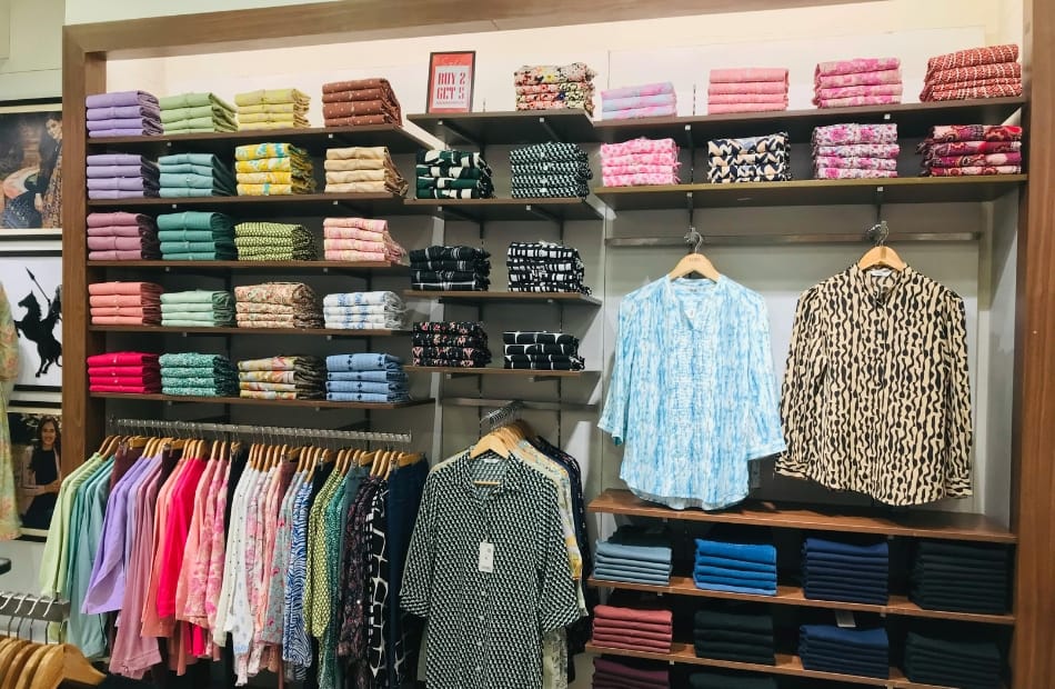 Display of stacked folded fabrics and some hanging tops in a clothing store, with various colors and patterns, including animal prints and pastel shades.