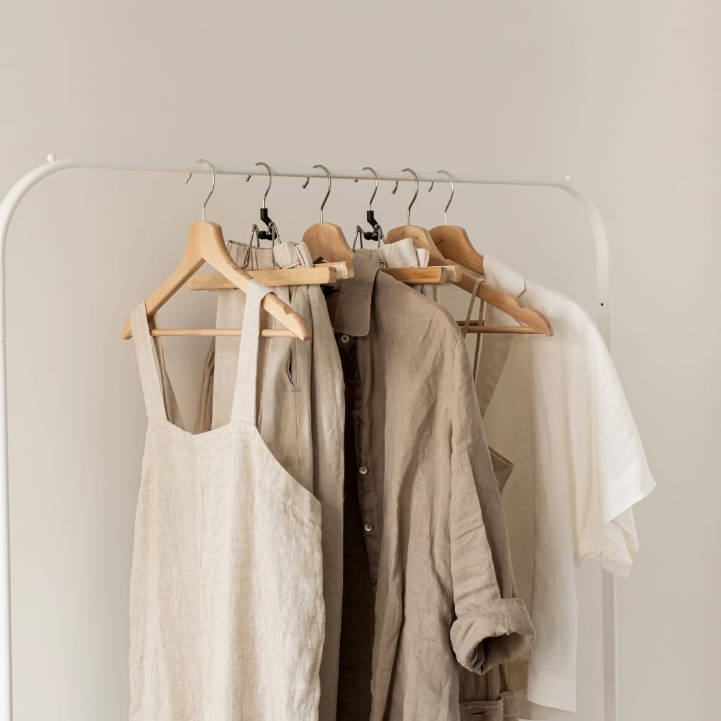 Several neutral-colored clothing items, including a linen dress, shirts, and a t-shirt, hanging on a white clothing rack against a plain background