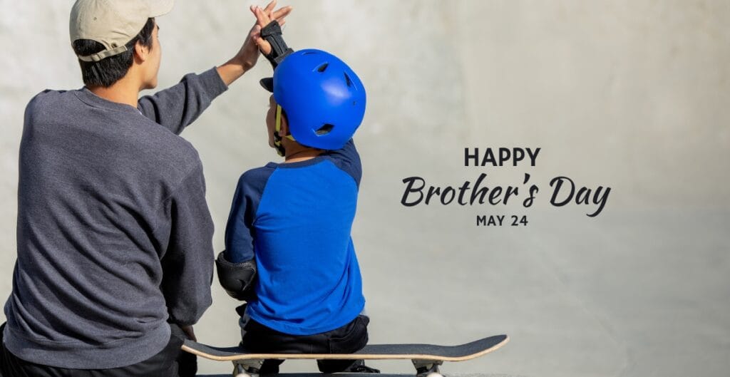 A young boy wearing a blue helmet sitting on a skateboard with an adult giving him a high-five, celebrating brotherhood.