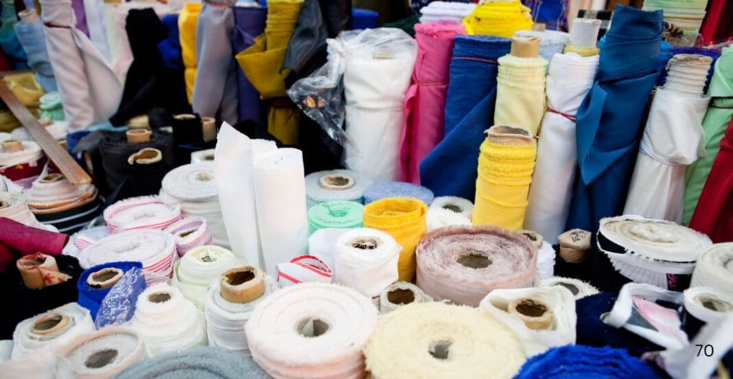 A colorful market stall displaying rolls of fabric in various colors and textures, with some fabric spools wrapped in plastic, set against a backdrop of hanging fabrics.