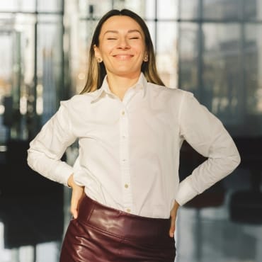 A woman smiling confidently, wearing a white button-up polyester shirt tucked into a burgundy pencil skirt, standing in a modern indoor setting.