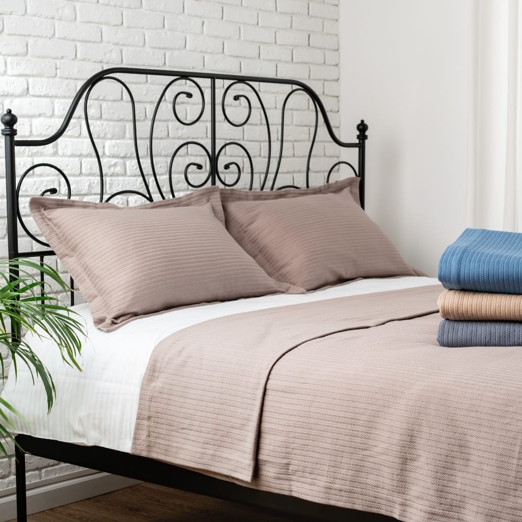 A neatly made bed with greyish-brown striped pillows and a matching bedspread, placed against a white brick wall. There is a stack of folded towels in shades of blue, beige, and gray on the right side of the bed, and a green houseplant with long leaves on the left side.