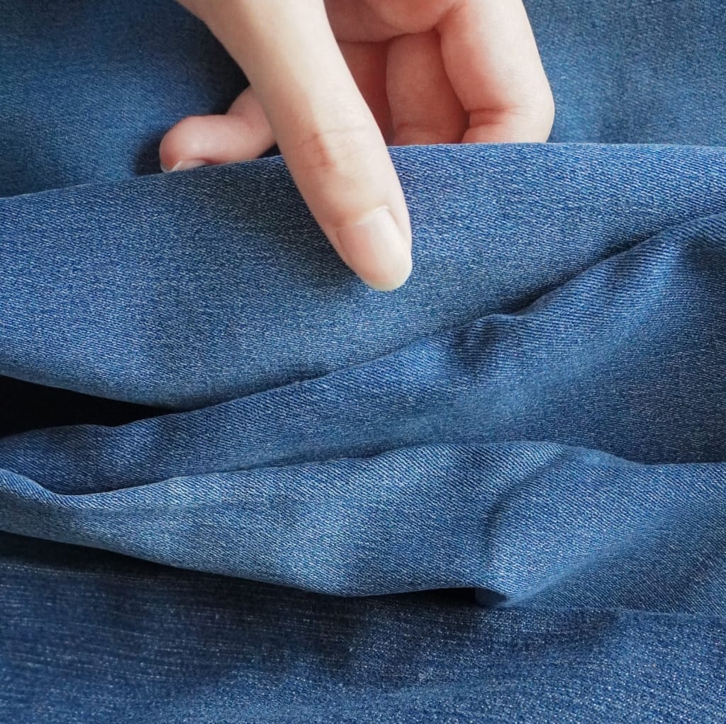 Close-up of a person's hand gently pinching a piece of blue denim cotton fabric, showing its texture and weave pattern.