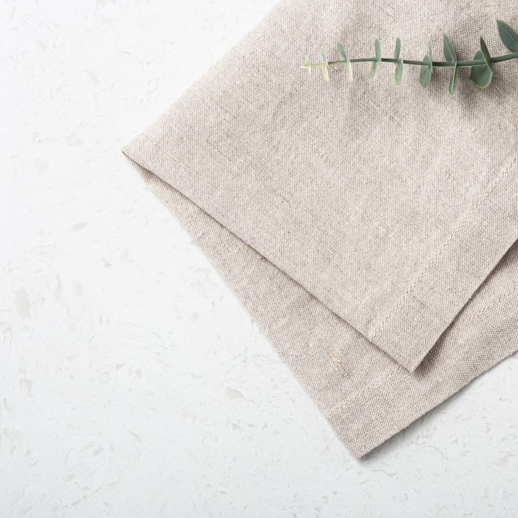 Stacked beige linen napkins with a sprig of eucalyptus on top, placed on a white textured surface.