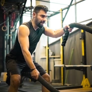 A man engaged in a high-intensity workout, gripping battle ropes in a gym, wearing a sleeveless athletic shirt made of polyester jersey fabric.