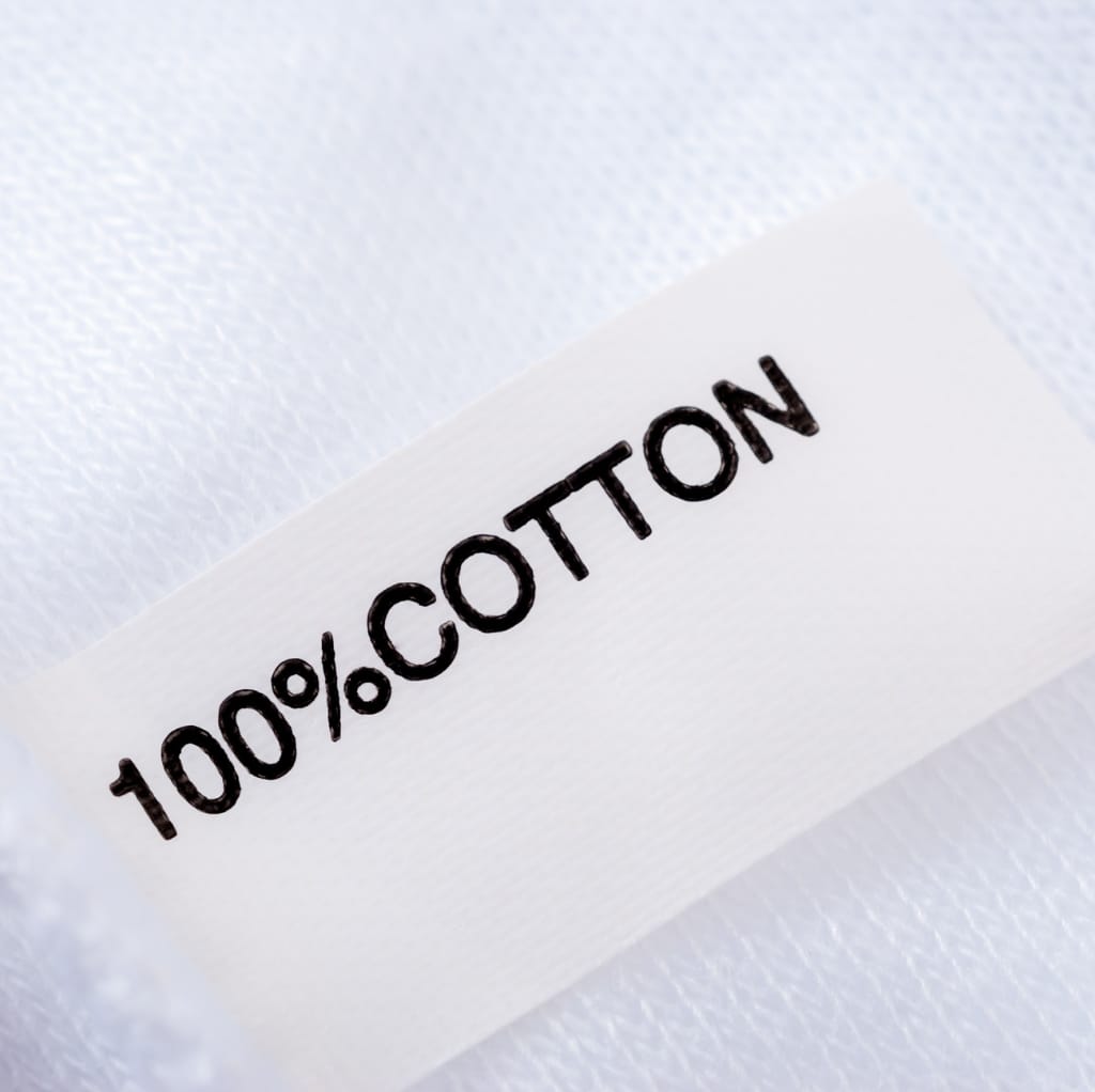 Close-up of a white fabric label with the words "100% COTTON" written in bold black letters, resting on a similar textured fabric background.