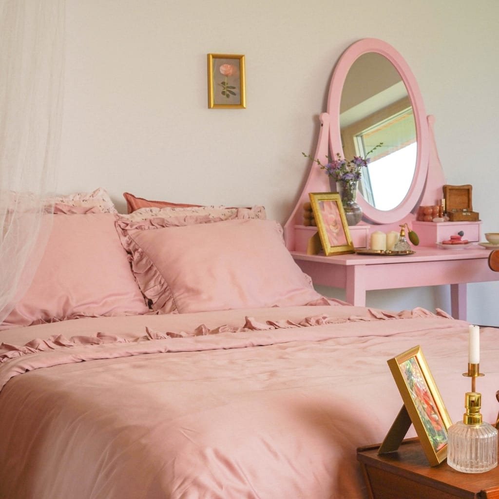A cozy bedroom with pink ruffled bedding, a pink vanity with a mirror, and decorative items, creating a soft and feminine atmosphere.