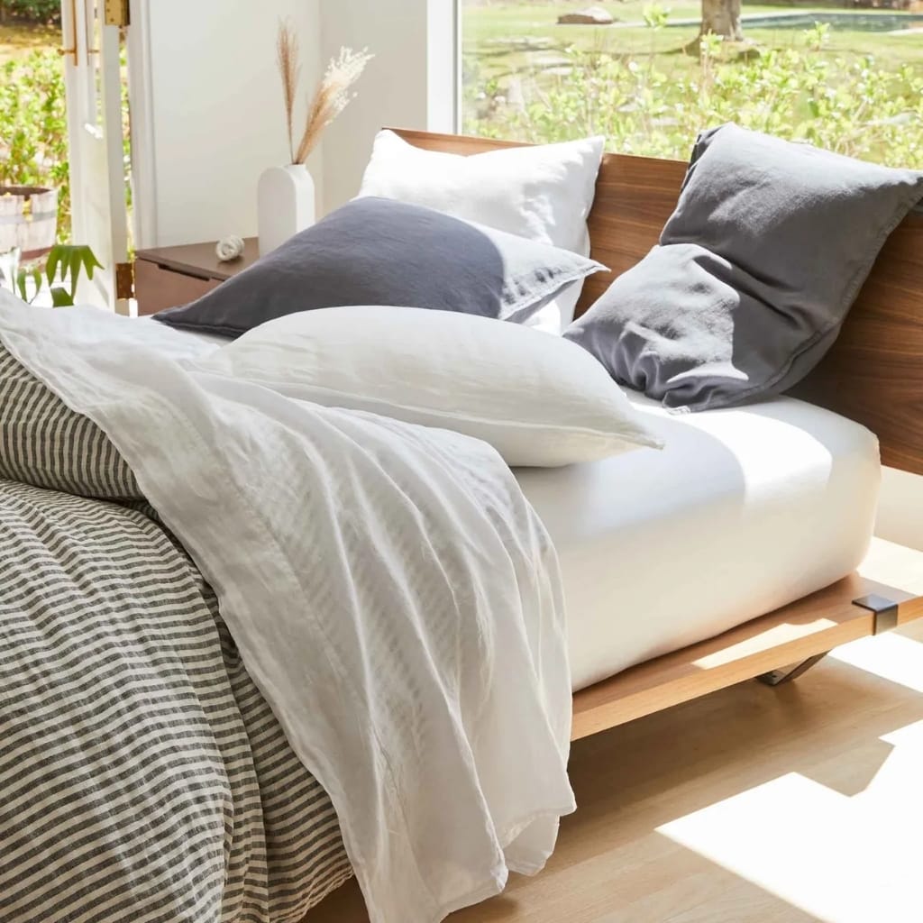 A cozy bedroom corner with a bed featuring a wooden headboard, dressed in white and grey bedding, near large windows letting in natural sunlight, with green outdoor scenery visible.