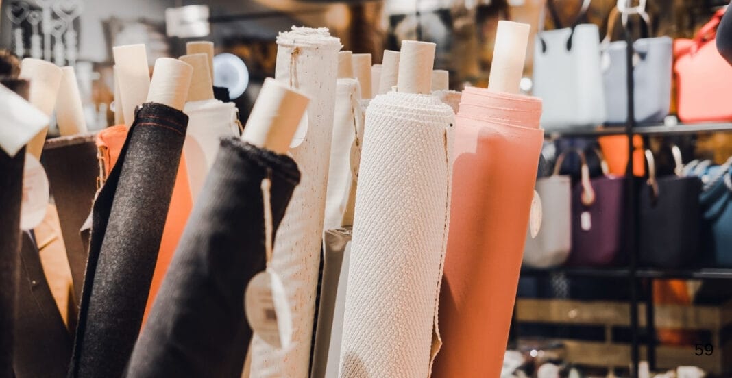 A display of various rolls of fabric, including cotton, in different colors such as black, white, peach, and beige, arranged upright on shelves in a fabric store.