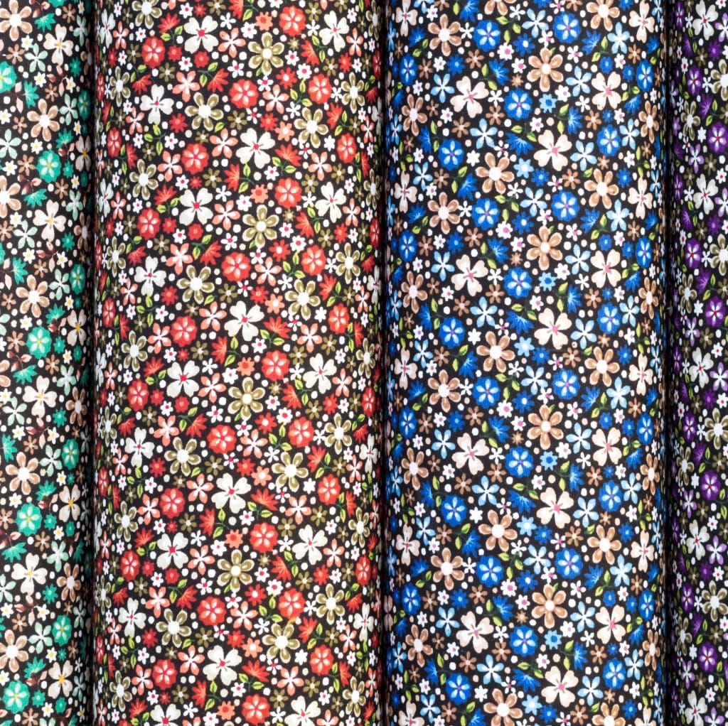Close-up image of multiple rolls of cotton fabric with floral patterns, arranged vertically. The fabrics feature various colorful flower designs on dark backgrounds.
