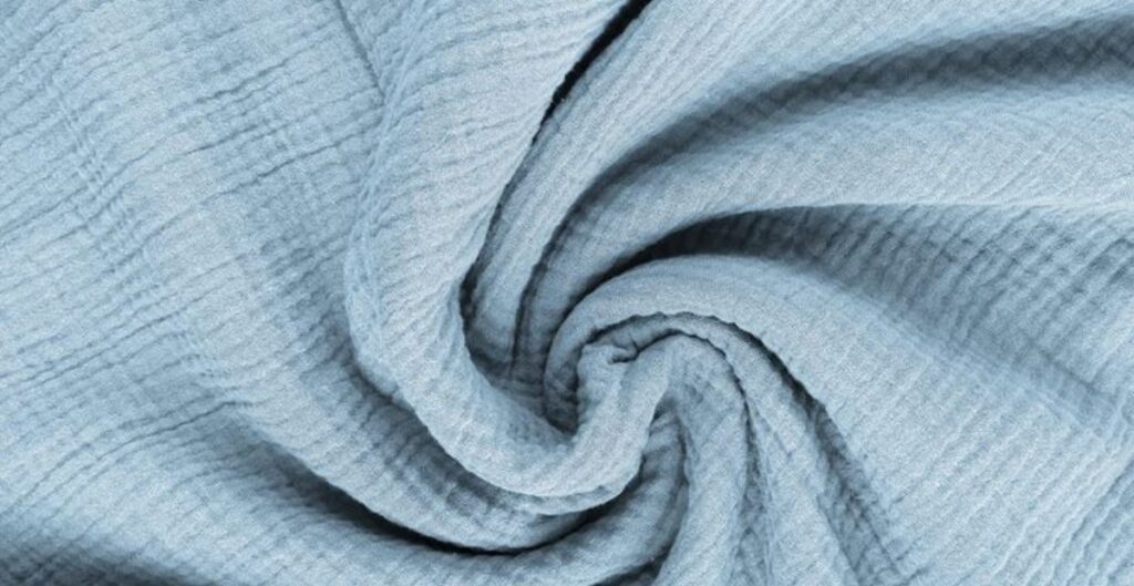 A close-up image of light blue cotton gauze fabric, twisted into a spiral. The fabric has a soft, airy, and textured appearance with a slightly wrinkled surface.