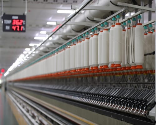 A textile manufacturing machine with white spools of thread or yarn arranged along it, with digital gauges displaying temperature readings. Top Global Companies