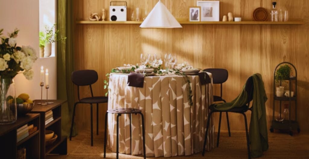 A cozy dining area featuring a round table draped with a beige and white patterned tablecloth, surrounded by four black chairs. The table is set with glassware and cutlery, and there are candles on a small side table to the left. The background has wooden paneled walls with a floating shelf holding framed photos and decorative items. Soft light fills the room, creating a warm atmosphere.