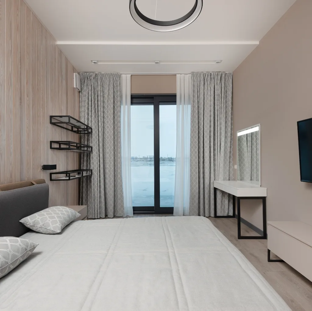 A cozy bedroom with a large bed, soft pillows, minimalist black wall shelves, a vanity with a mirror, light-colored curtains, and a view of a waterfront through a glass door. Cotton Textiles Made in USA