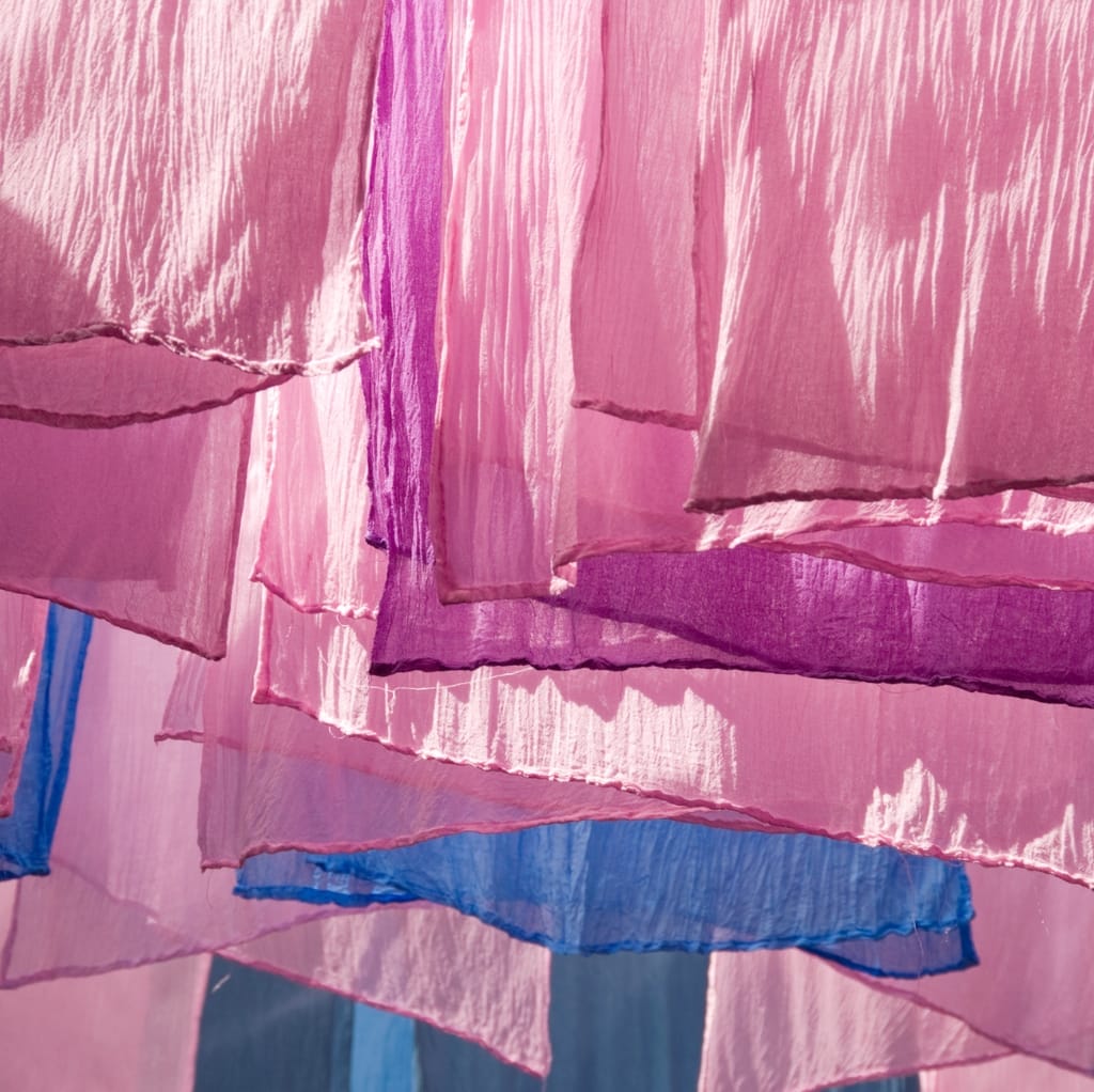 Colorful strips of lightweight cotton fabric in shades of pink, purple, and blue hanging, with sunlight shining through, highlighting their textures.