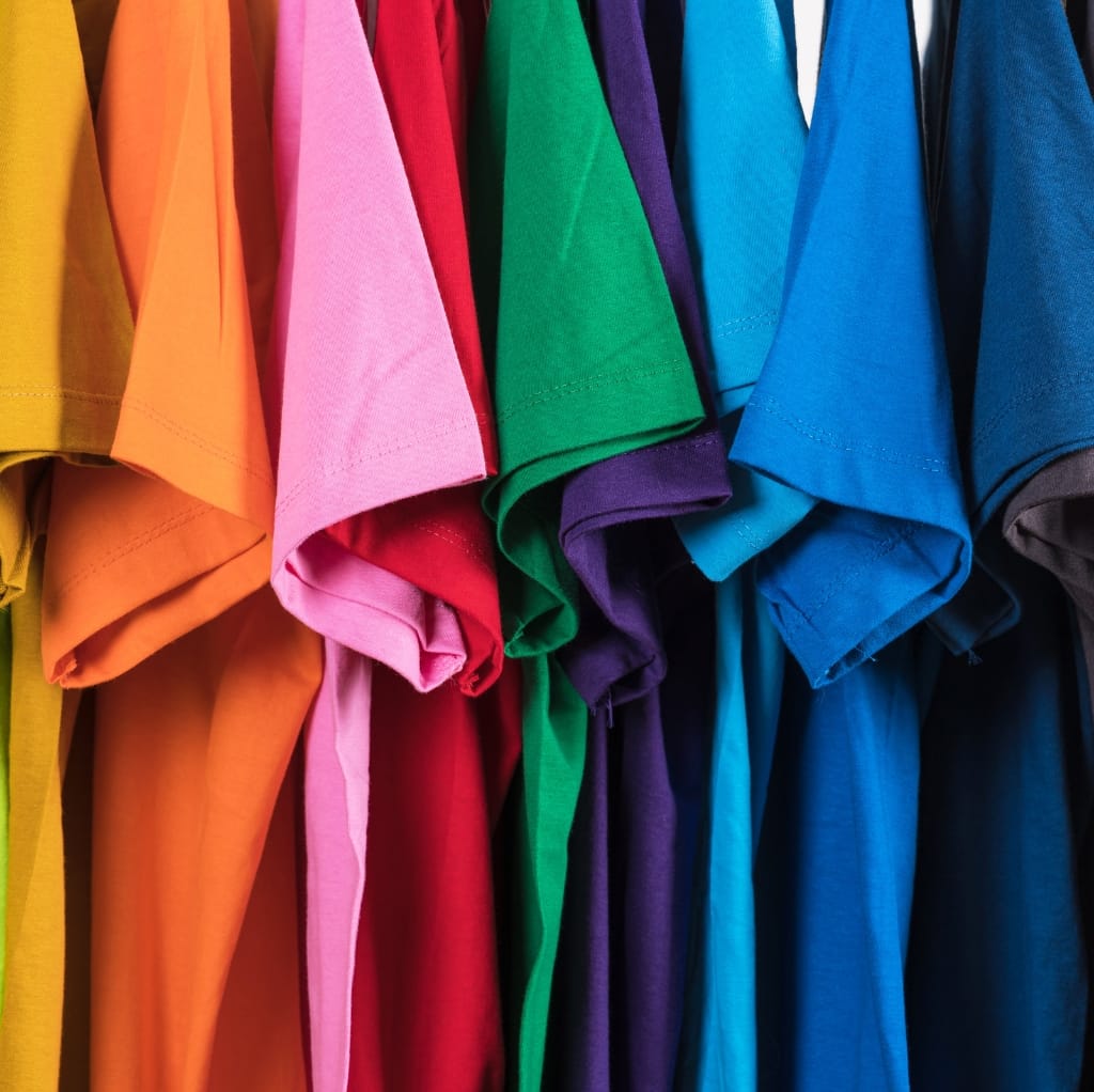 A collection of colorful t-shirts hanging together, featuring various shades such as yellow, orange, pink, red, green, purple, blue, and dark blue.