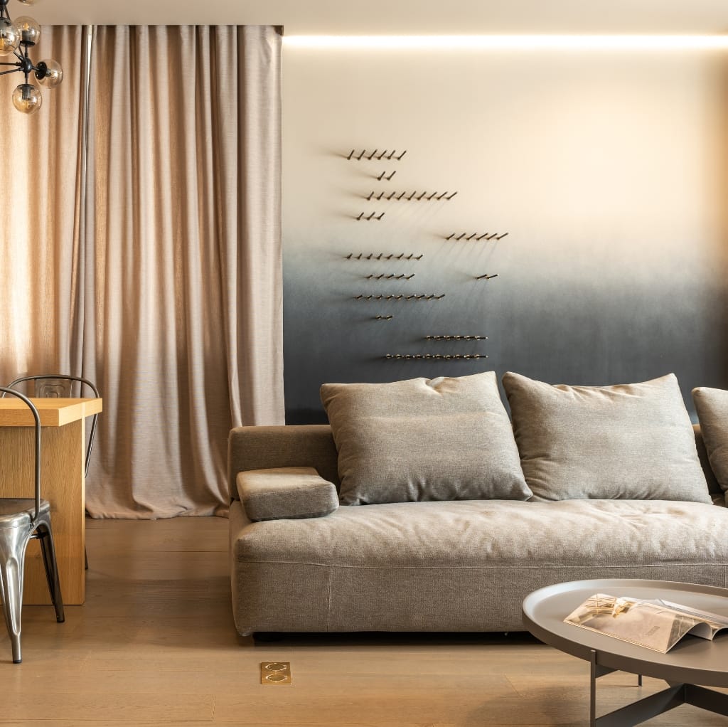 A modern living room corner featuring a beige fabric sofa with matching cushions, a round white coffee table with a magazine, a wooden dining table with metal chairs, beige curtains, and a unique wall art installation that resembles a formation of airplanes.