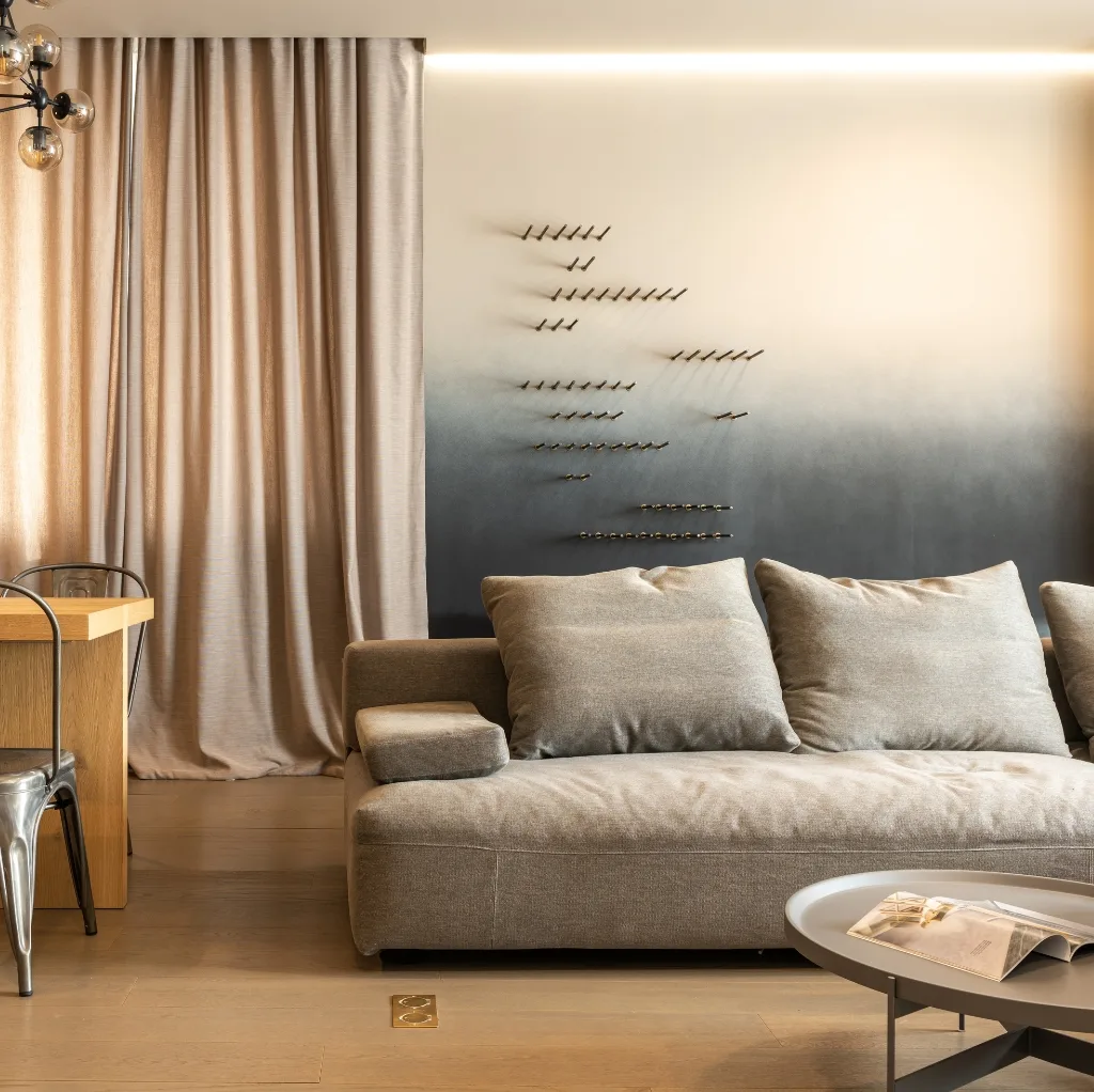 A modern living room corner featuring a beige fabric sofa with matching cushions, a round white coffee table with a magazine, a wooden dining table with metal chairs, beige curtains, and a unique wall art installation that resembles a formation of airplanes.