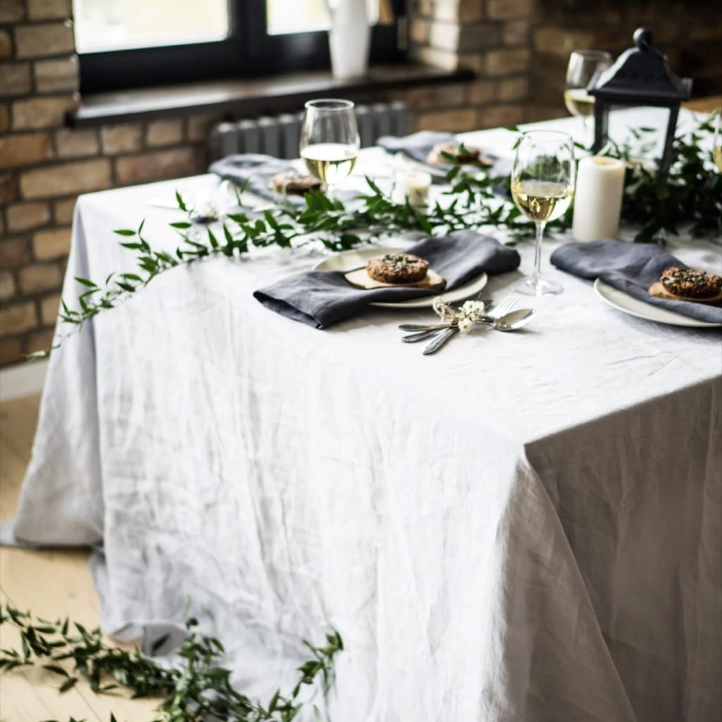 A beautifully set dining table with a white tablecloth, decorated with green foliage, white candles, and a lantern. The table is set with black napkins, plates, and wine glasses filled with white wine, ready for a meal in a rustic, cozy setting with a brick wall in the background.