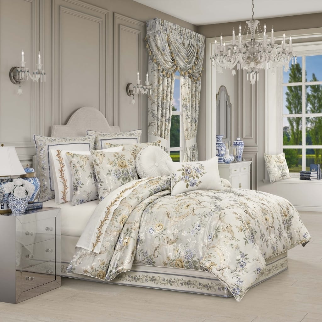 A luxurious bedroom with elegant, light-colored furnishings. The bed has a floral-patterned duvet and multiple decorative pillows. The room features a classic design with gray paneled walls, a white headboard, a chandelier, wall sconces, and draped curtains. Large windows reveal a lush green outdoor scene.