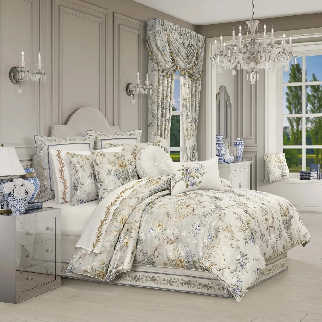 A luxurious bedroom with elegant, light-colored furnishings. The bed has a floral-patterned duvet and multiple decorative pillows. The room features a classic design with gray paneled walls, a white headboard, a chandelier, wall sconces, and draped curtains. Large windows reveal a lush green outdoor scene.