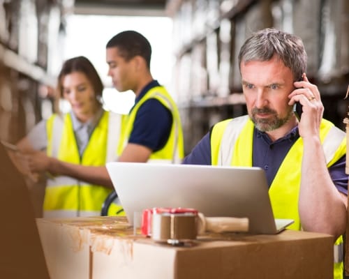 A man wearing a yellow safety vest working on a laptop while talking on the phone in a warehouse, with two other workers in safety vests in the background consulting a tablet.