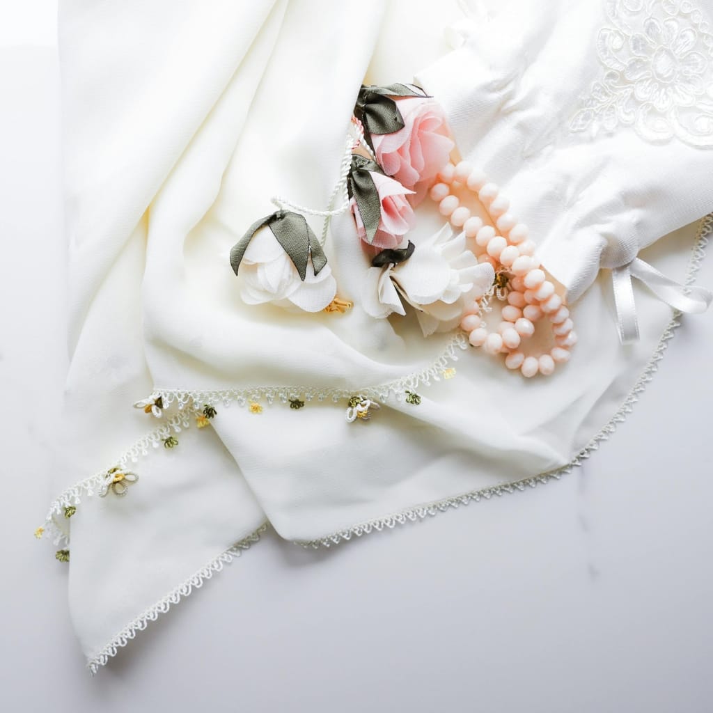 A delicate white fabric with embroidered details, adorned with pink and white fabric flowers, black ribbon bows, a string of pink beads, and a lace-trimmed piece of cloth.
