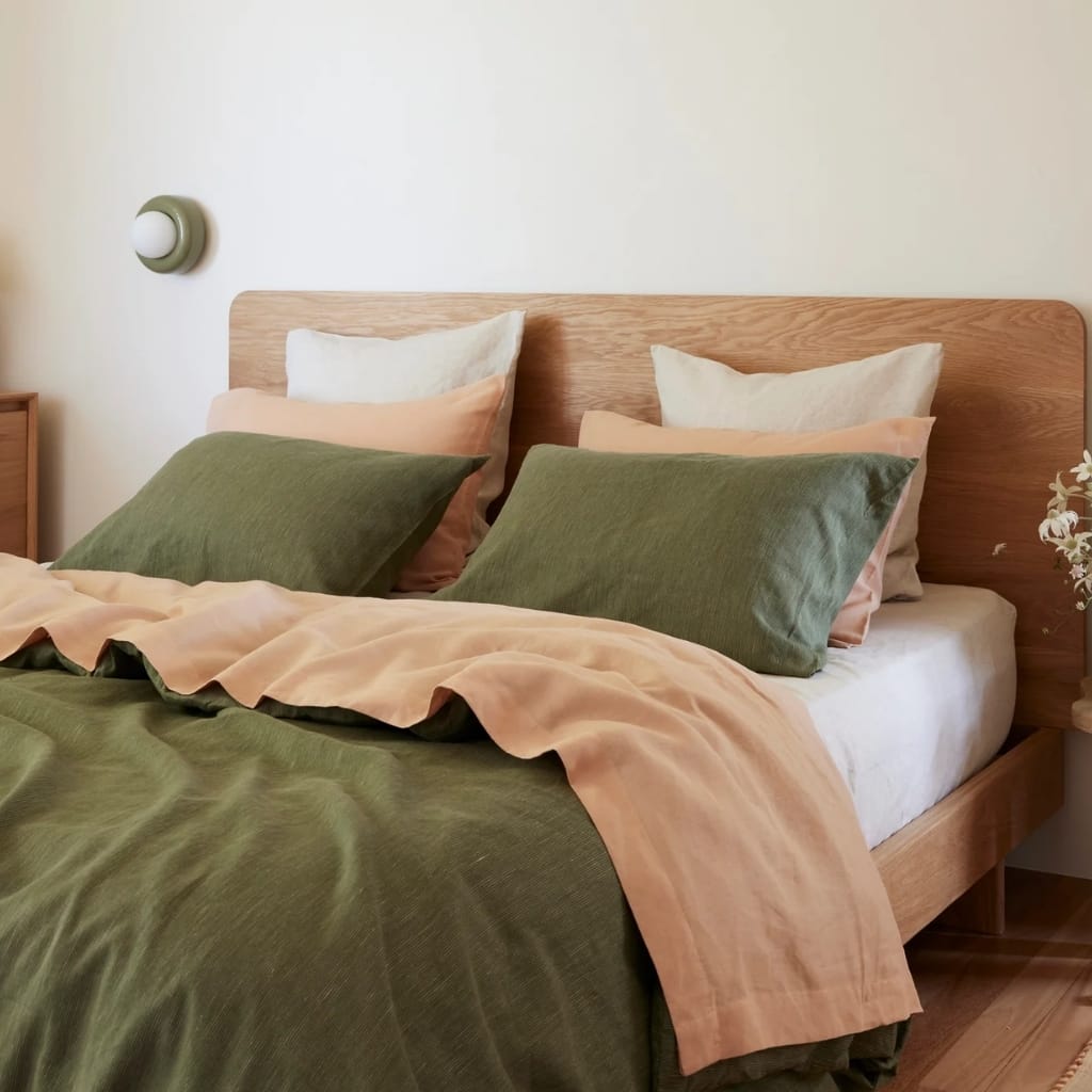 A neatly made bed with a natural wooden headboard, featuring four pillows (two green and two beige) and a color-blocked duvet cover in shades of them. The bed is located in a cozy, minimalistic room with a small wooden bedside table on the left and a modern wall-mounted light fixture on the wall above.