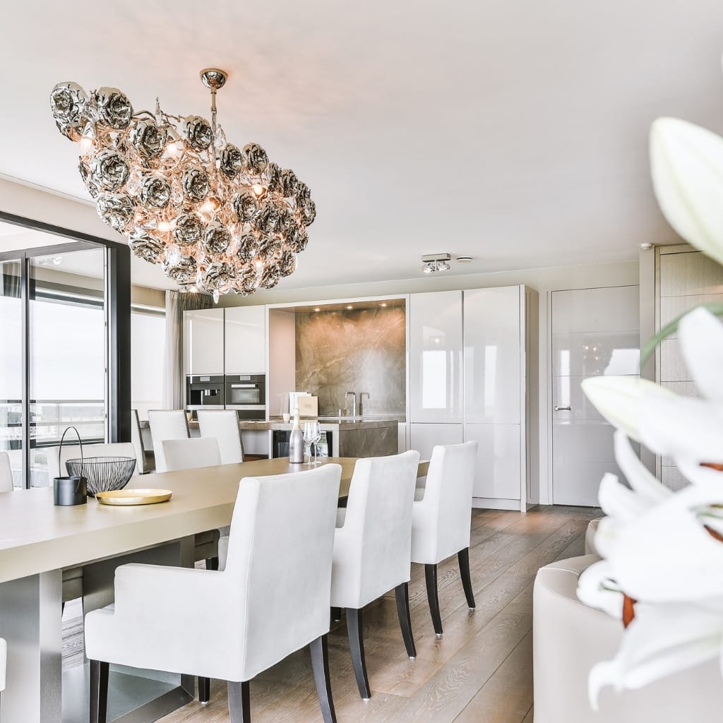 A modern, minimalist dining area featuring a large light-colored table with white upholstered chairs, a contemporary chandelier, and a bright open kitchen with sliding glass doors and natural light.