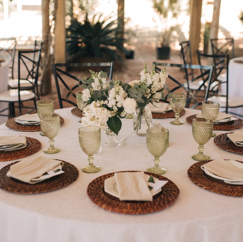 A round table set for an elegant event, featuring a white tablecloth, woven placemats, neatly folded cream napkins, vintage-style green glass goblets, and a central floral centerpiece of white flowers and greenery in a clear glass vase. The background reveals a charming outdoor setting with natural light and wooden chairs. Bulk Cotton Fabric