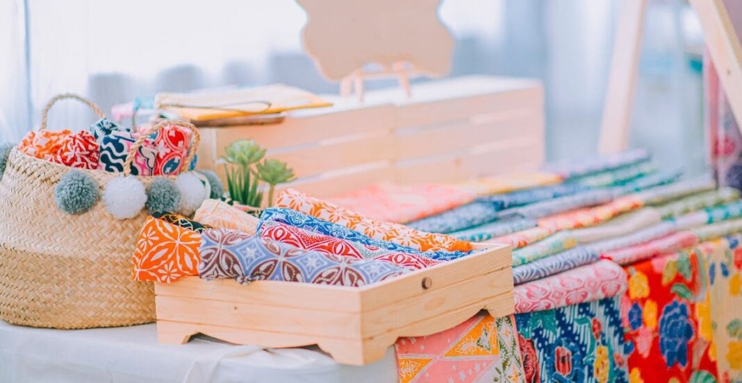 A display table with colorful patterned textiles and accessories, including a woven basket with pom-pom decorations, set up in a bright, airy space.