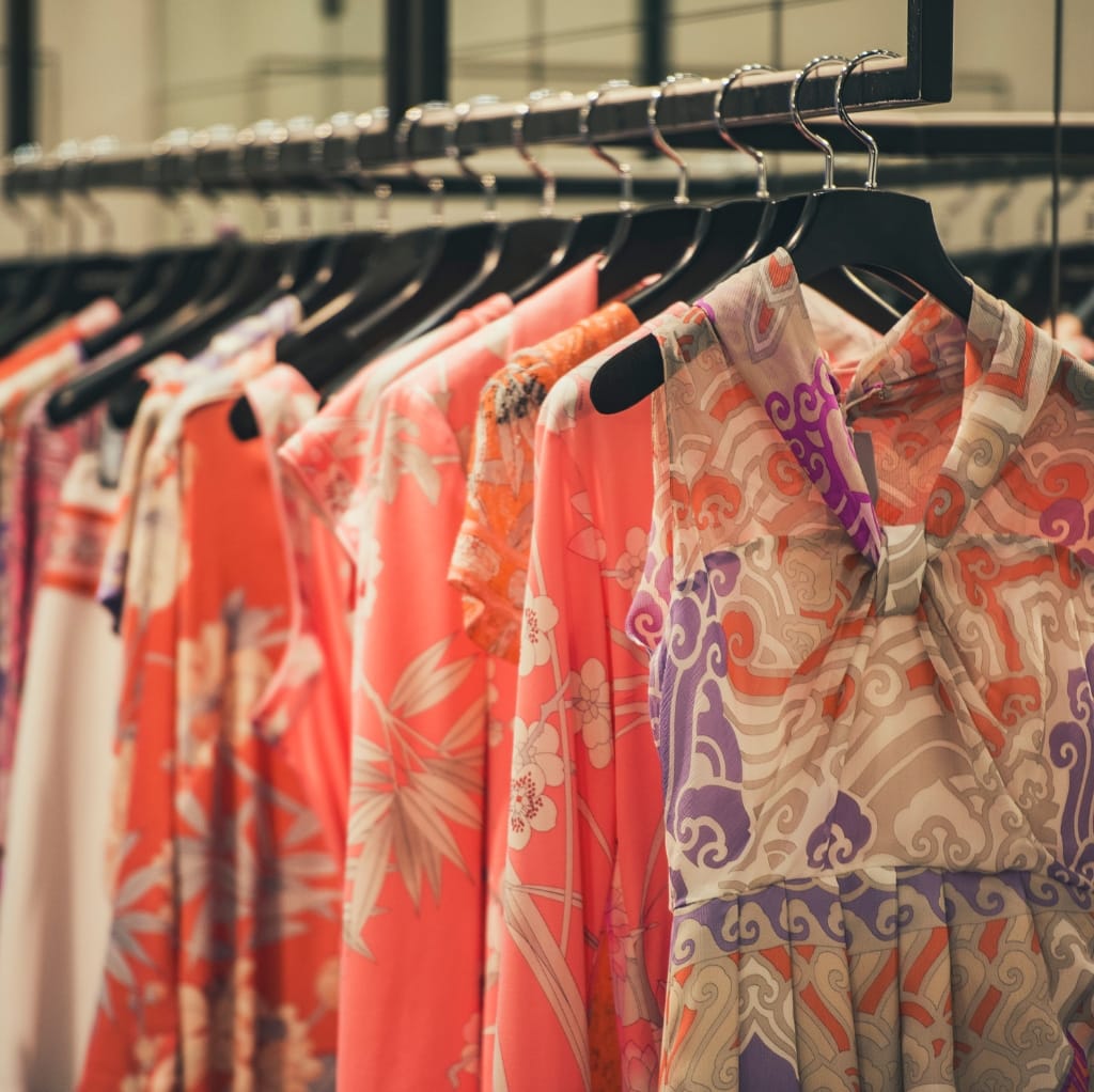A row of colorful, patterned cotton shirts and dresses hanging on black hangers across a metal clothing rack, showcasing vibrant fall fashion with floral and abstract designs.