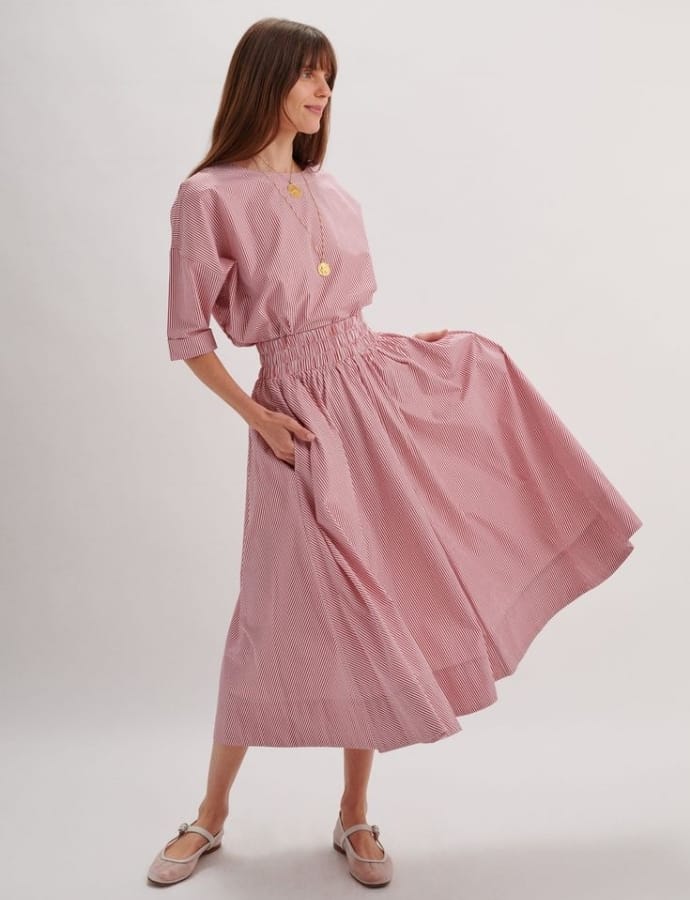 A woman in a pink and white striped dress, holding the hem of her skirt, standing against a plain white background.