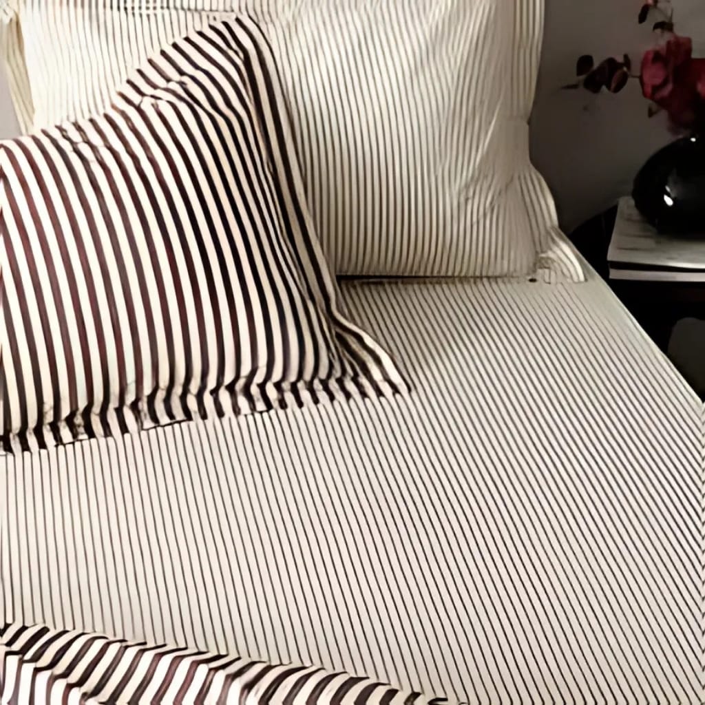 A close-up of a bed with white and black vertically striped bed sheets and pillowcases. The bed is next to a dark-colored nightstand with a black vase containing pink flowers and some books or magazines.