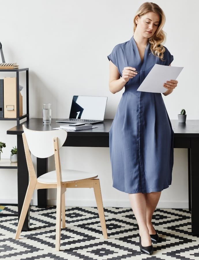 A woman standing in a modern office space, wearing a chic blue cotton dress with black heels, holding some papers while reviewing them.