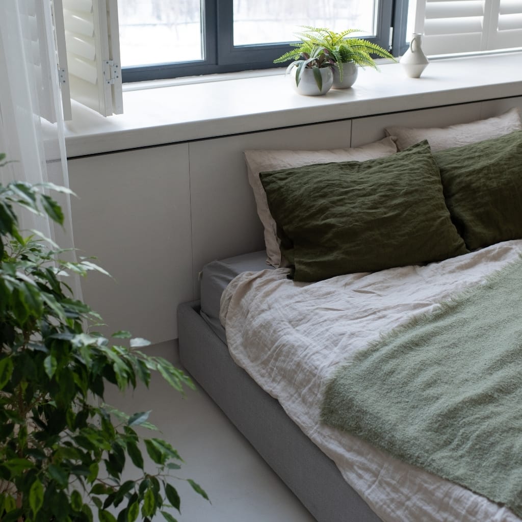 A cozy bedroom corner featuring a bed with beige and olive green pillows, a white linen blanket, and a white bed frame, with a wide window sill decorated with potted plants, and a large green plant visible in the foreground on the left.