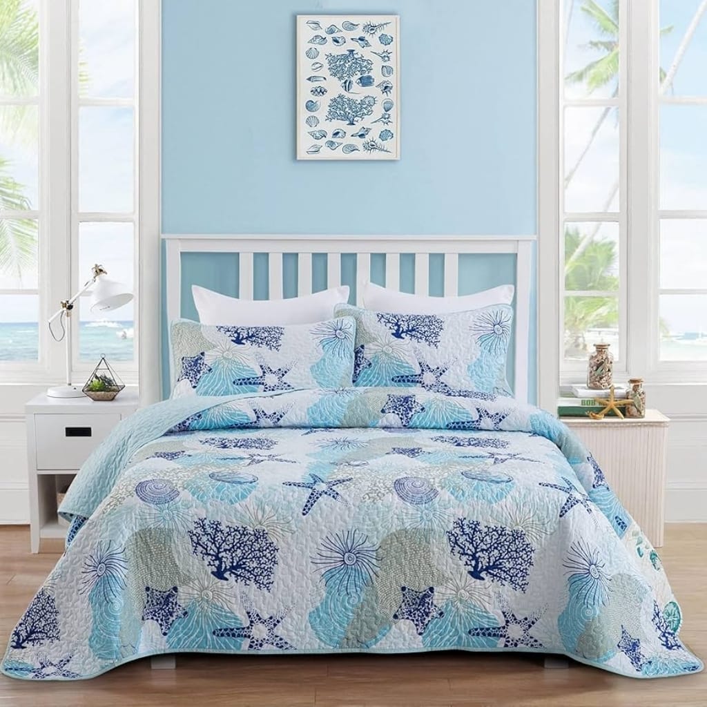 A cozy bedroom with a light blue wall and large windows showing a view of the ocean and palm trees outside. The bed has a white wooden headboard and is covered with a quilt and pillows featuring a marine-inspired pattern of starfish, sea shells, and coral in various shades of blue, teal, and cream. On each side of the bed are white nightstands, one with a modern white lamp and the other with small decorative jars, books, and sea-themed decor. A framed art print of various fish hangs on the wall above the bed.
