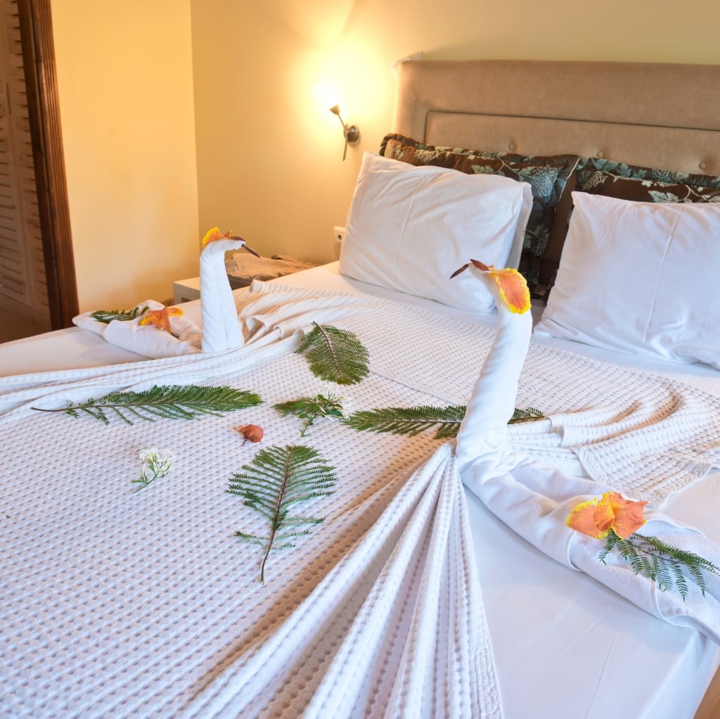 A neatly made hotel bed decorated with towel art in the shape of two swans, facing each other. The towels are adorned with flower petals and green leaves, creating a romantic and elegant presentation.