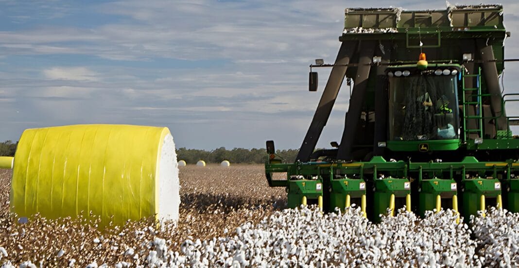 A green John Deere cotton picker machine harvesting cotton in a field with large yellow and white cotton bales in the background under a partly cloudy sky.