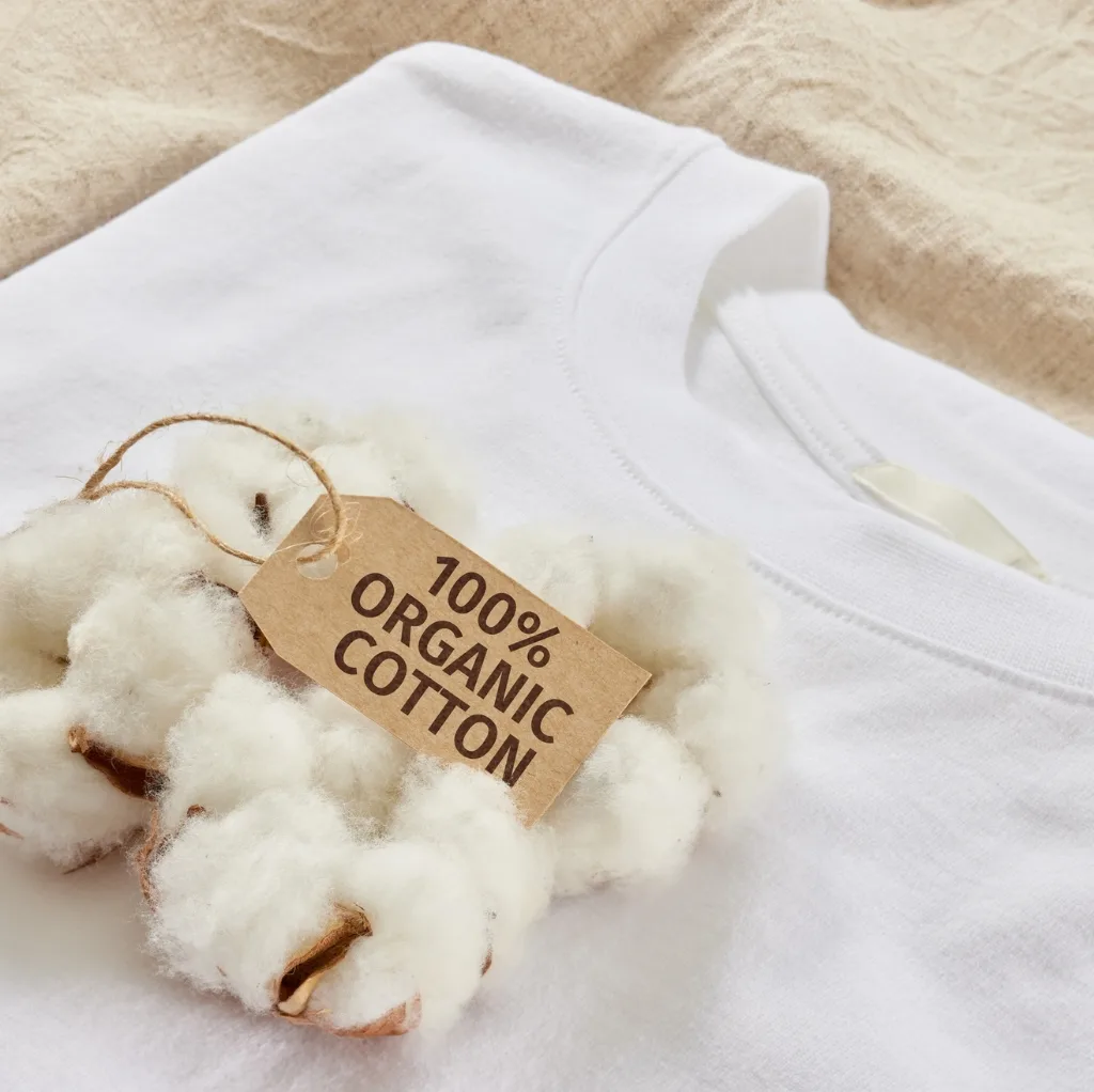 Close-up of a white cotton T-shirt with a tag reading "100% Organic Cotton" placed on it. In the foreground, a small cluster of fluffy cotton bolls.