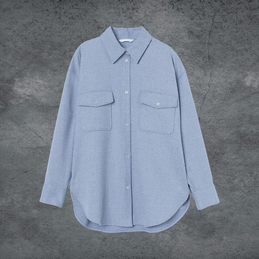 A light blue, long-sleeved button-up shirt with two chest pockets, laid flat against a textured gray background.