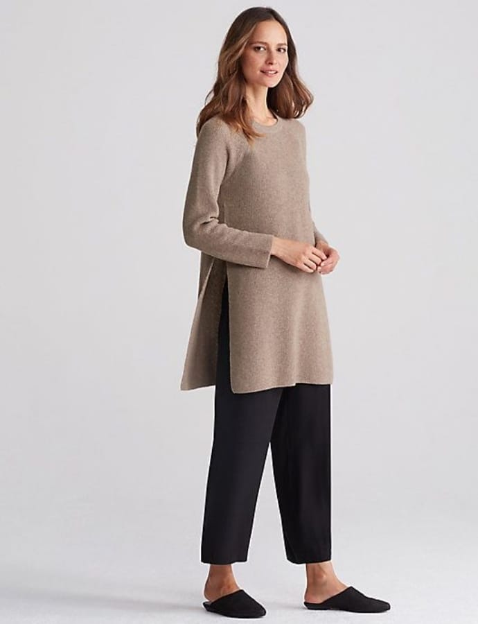 A woman wearing a beige long-sleeve tunic paired with black cropped trousers and black slip-on shoes, standing against a plain light grey background.