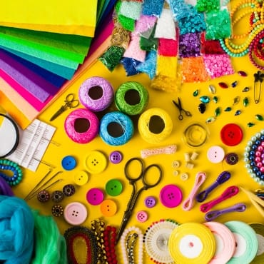 Bright and colorful assortment of craft supplies, including yarn, buttons, scissors, beads, and fabric, set against a yellow background, with various textures and embellishments.