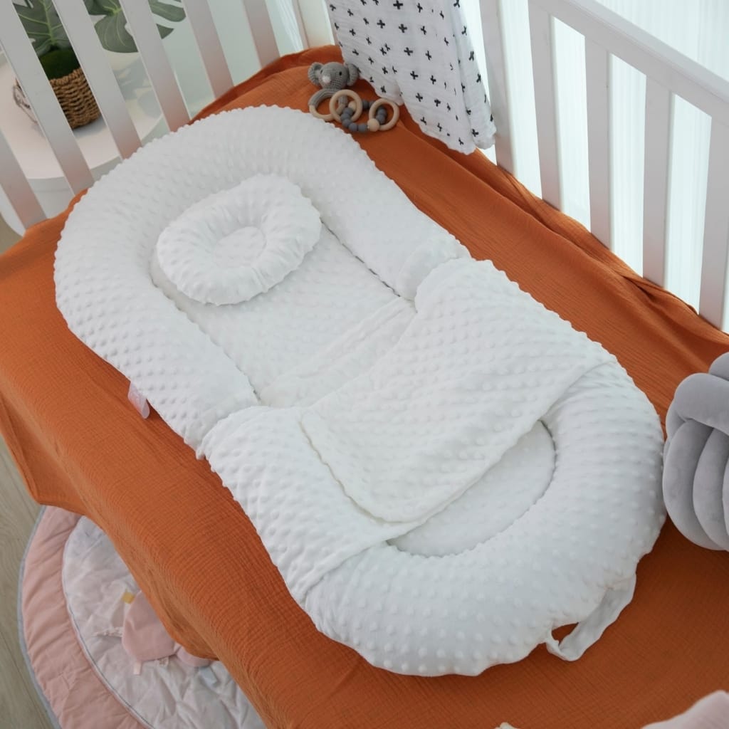 A white minky textured baby nest or lounger with a matching bumper and circular pillow inside, placed on a bed with an orange sheet. The baby nest has soft, raised edges and is positioned next to a white crib, with a small grey stuffed mouse and a few teething rings nearby. Light is coming through white blinds and patterned curtains in the background.