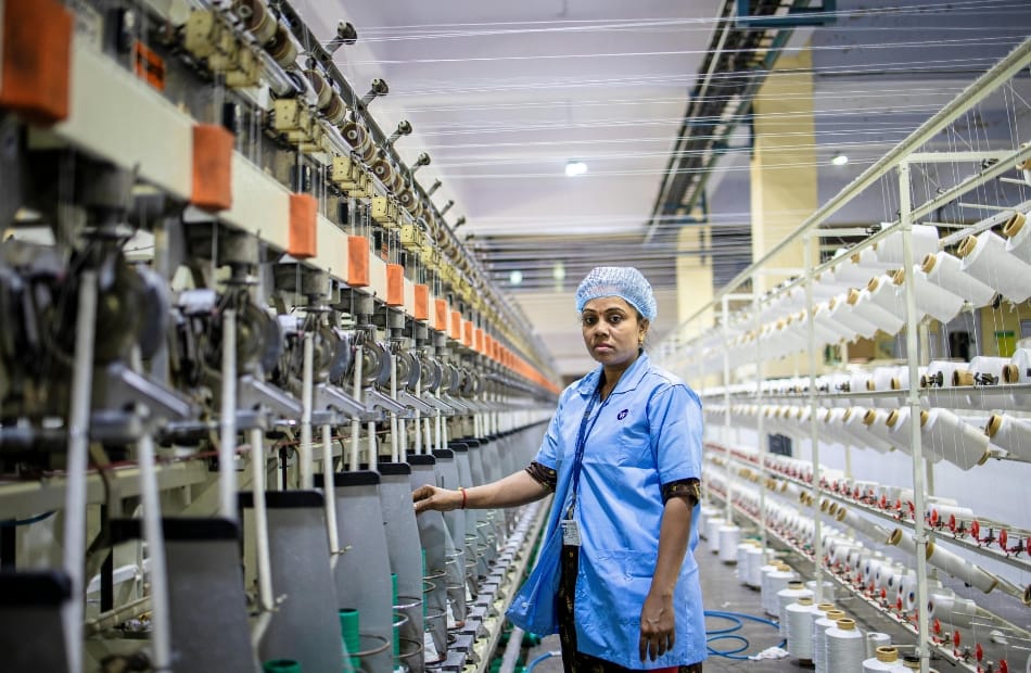 A woman wearing a blue coat and hair net standing in a busy textile manufacturing mill, with rows of spinning machines and spools of white cotton yarn around her. What Companies Produce Cotton