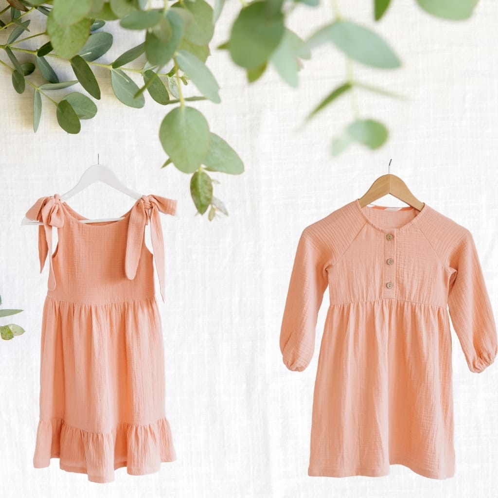 Two peach-colored dresses hanging on wooden and white hangers against a light, textured background, with green leaves partially visible in the upper part of the image.