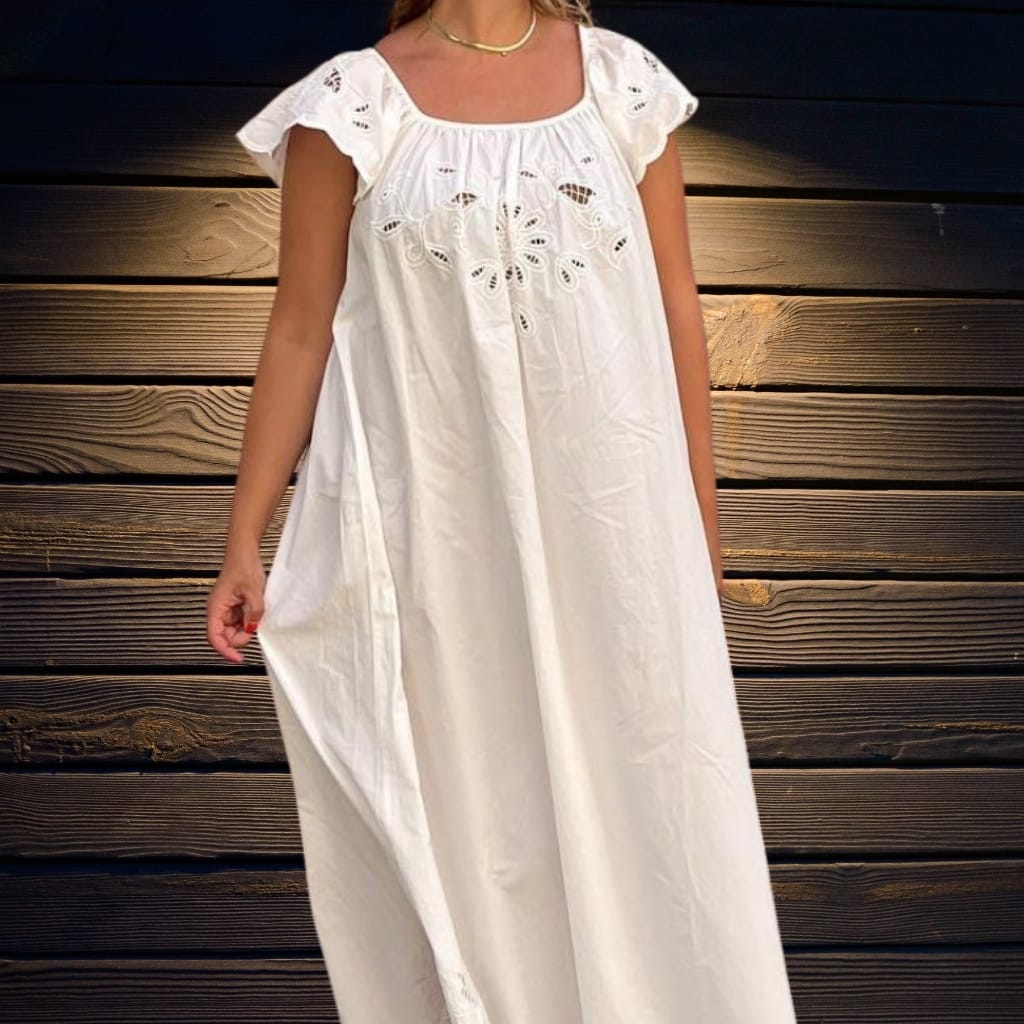 A woman wearing a white, embroidered, loose-fitting dress with short, ruffled sleeves standing in front of a dark wooden wall.
