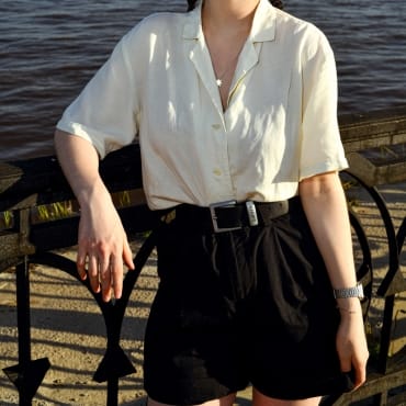 A person leaning on a black railing near water, wearing a layered outfit with a button-down shirt over a crop top.