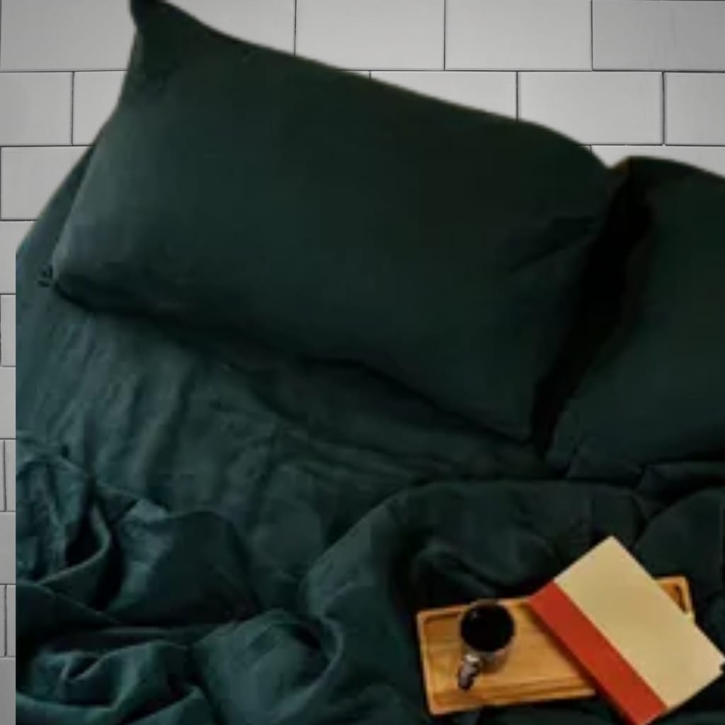 A dark green pillow resting on a similarly dark green bedsheet, with a small wooden tray holding a black cup and an orange notebook, placed on a bed against a tiled wall background.