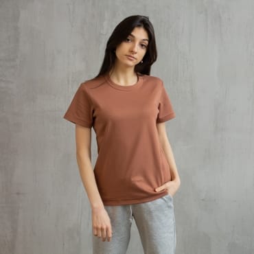 A young woman with long dark hair wearing a loose-fitting, short-sleeved brown polyester t-shirt and light gray sweatpants standing against a plain gray wall.